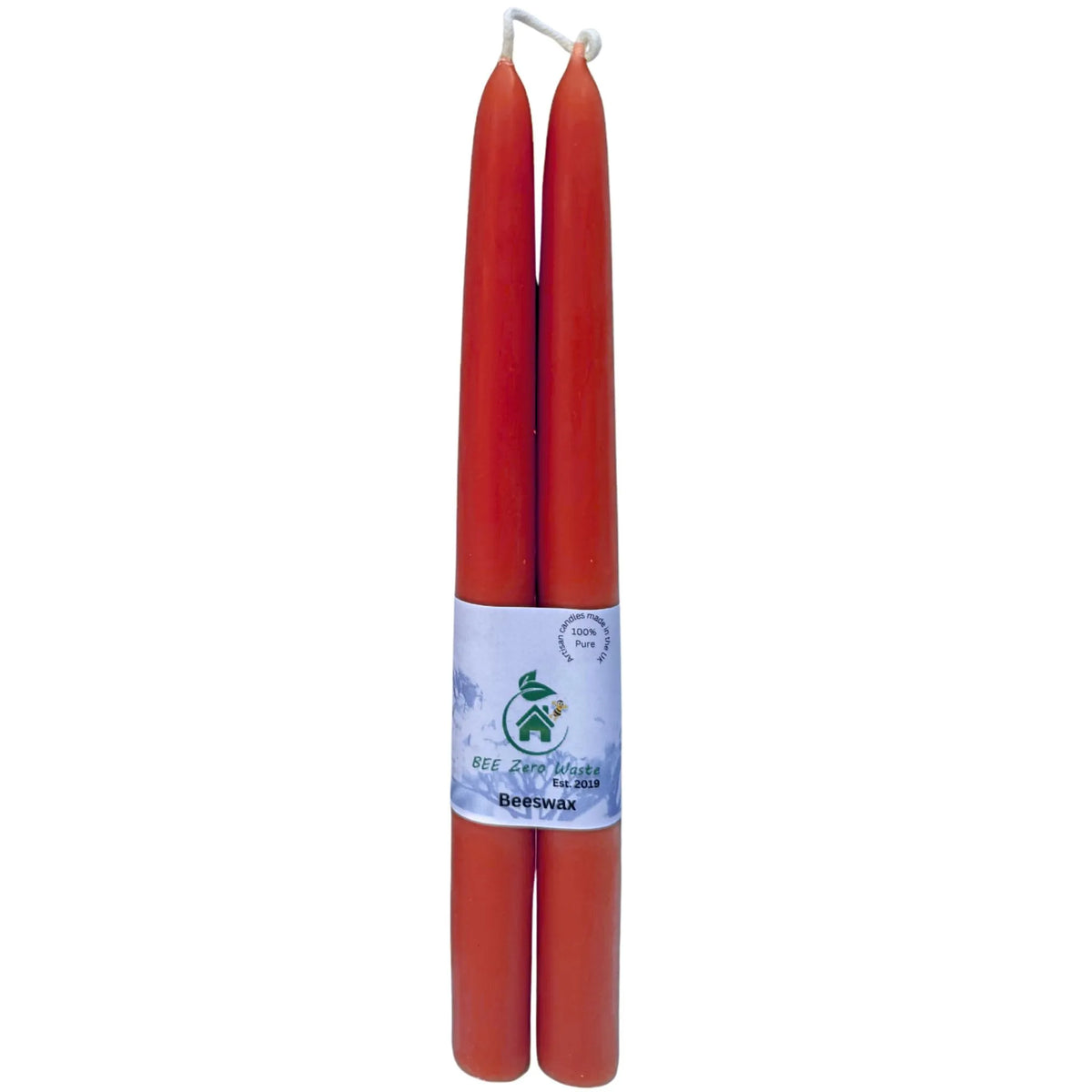 Two red taper candles standing upright side by side, with a white paper label wrapped around the middle. The label features a green logo and text, including 'BEE ZWAKI' and 'TAPER CANDLE'.