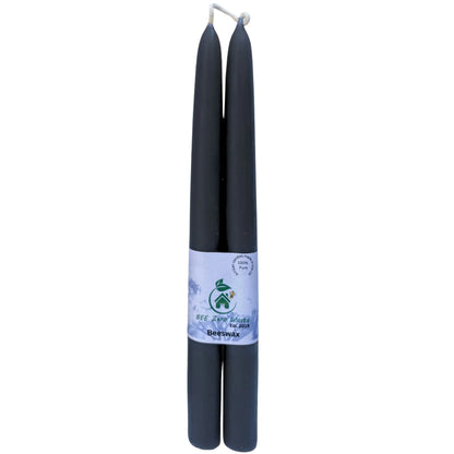 Two black beeswax taper candles wrapped together with a white paper band featuring a green logo and text, set against a plain white background.