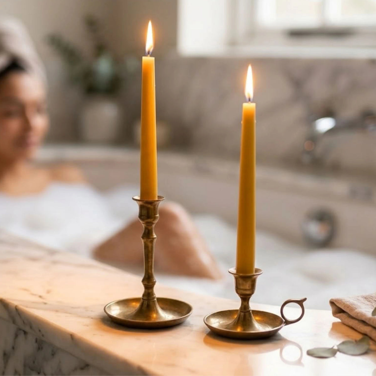 Two lit taper candles in brass candlesticks on a marble surface, with a blurred person sitting in a bathtub in the background.